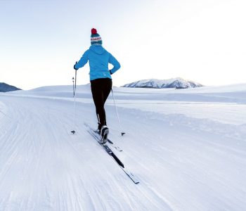 Langläuferin mit blauer Jacke und Mütze auf Skipiste, am Horizont sind Berge zu sehen. 
