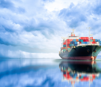 Ein Frachtschiff, vollgeladen mit Containern, spiegelt sich im Meer vor einem wolkenverhangenen Himmel. 