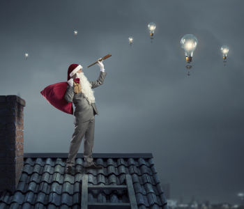 Weihnachtsmann im Anzug, mit Bart, Mütze und Geschenkesack, steht auf einem Dach und schaut mit einem Fernrohr in den Nachthimmel, in dem Glühbirnen schweben, die eine Idee symbolisieren.