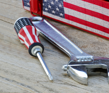 Schraubenschlüssel und Schraubendreher mit USA-Flaggenaufdruck im Griff auf einem Holzboden und vor einer Kiste mit USA-Flaggenaufdruck.