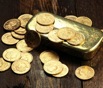 Ein gllänzender Goldbarren und einige verteilte Goldmünzen liegen auf einem dunklen Holztisch.